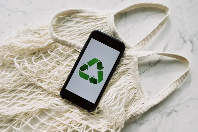 Top view of mobile phone with green recycling emblem on screen composed on white mesh bag on marble table in room. Retail sustainability claims