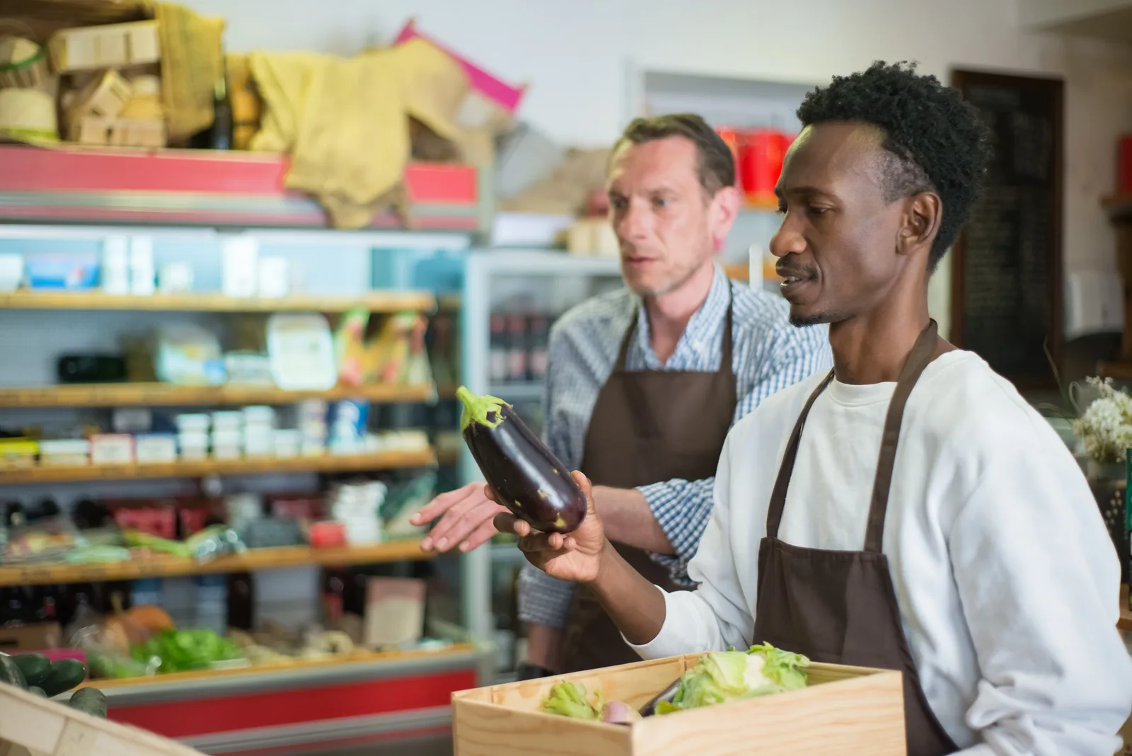 Two workers organizing vegetables in a grocery store, focusing on fresh eggplants.